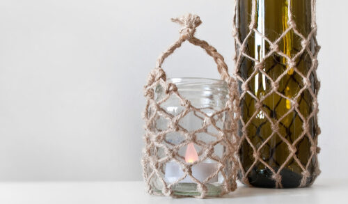 A clear glass jar wrapped in rope with a tea light next to a taller, green bottle.