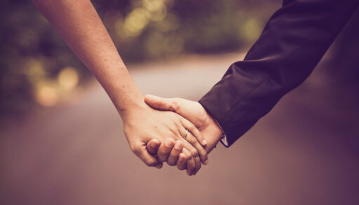 Close-up of a bride and groom holding hands with a pathway and trees in the background.