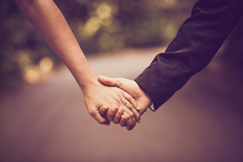 Close-up of a bride and groom holding hands with a pathway and trees in the background.