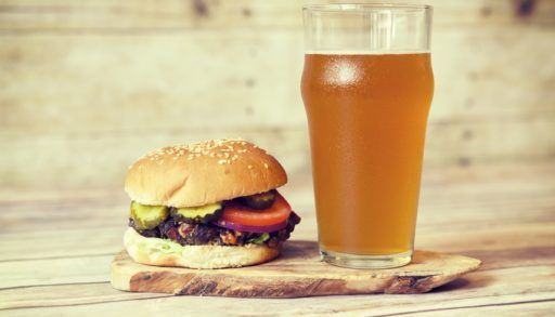 Glass of amber-coloured beer next to a hamburger topped with pickles and tomato.