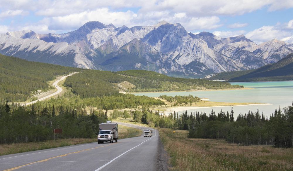 Truck pulling an RV on a road surrounded by mountains and near a lake on the Kootenay Plains, Alberta.