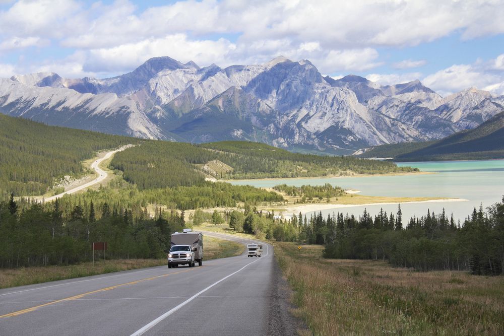 Truck pulling an RV on a road surrounded by mountains and near a lake on the Kootenay Plains, Alberta.