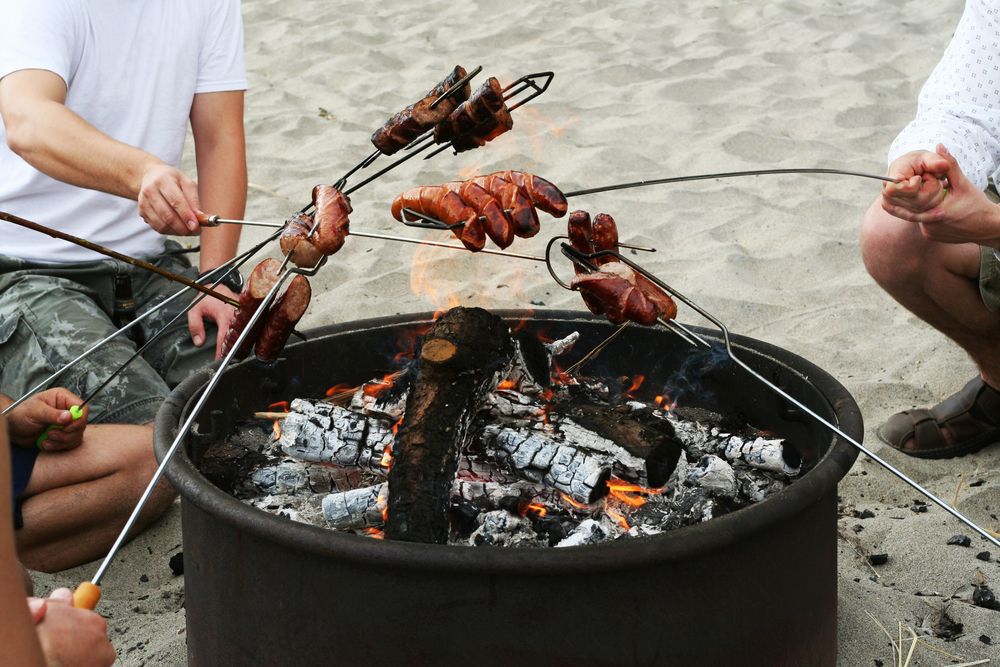 Group of people cooking sausages over a campfire on the beach.
