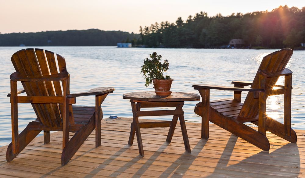 Two wooden Muskoka chairs on a dock next to a lake at sunset.
