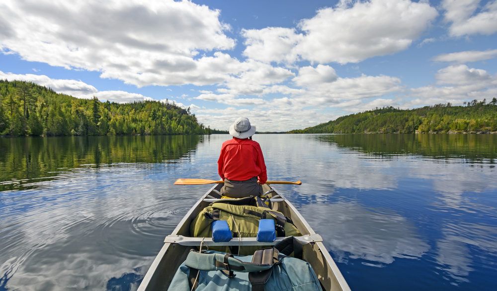 Canoeist sitting in the front of a canoe on a lake with green trees on either side.