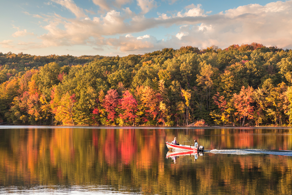 A motor boat on a still lake with red and green trees reflected in its surface.