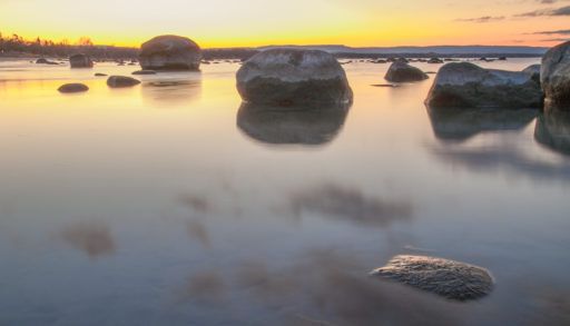 Sunset over a still Lake Huron with large rocks standing in the water.