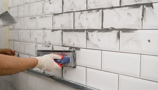 Person wearing work gloves applying grey grout to white wall tiles.