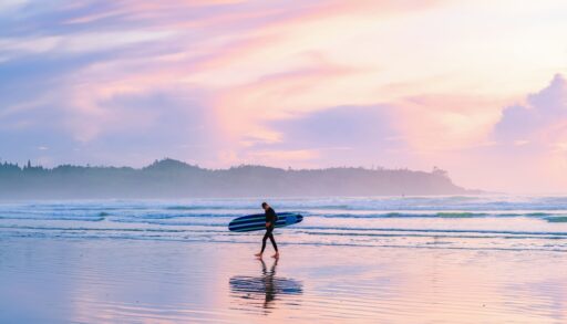 A person walking along Tofino Beach, B.C. with a surfboard at sunset.