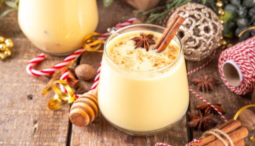 A glass of eggnog with a cinnamon stick on a table with holiday decorations.
