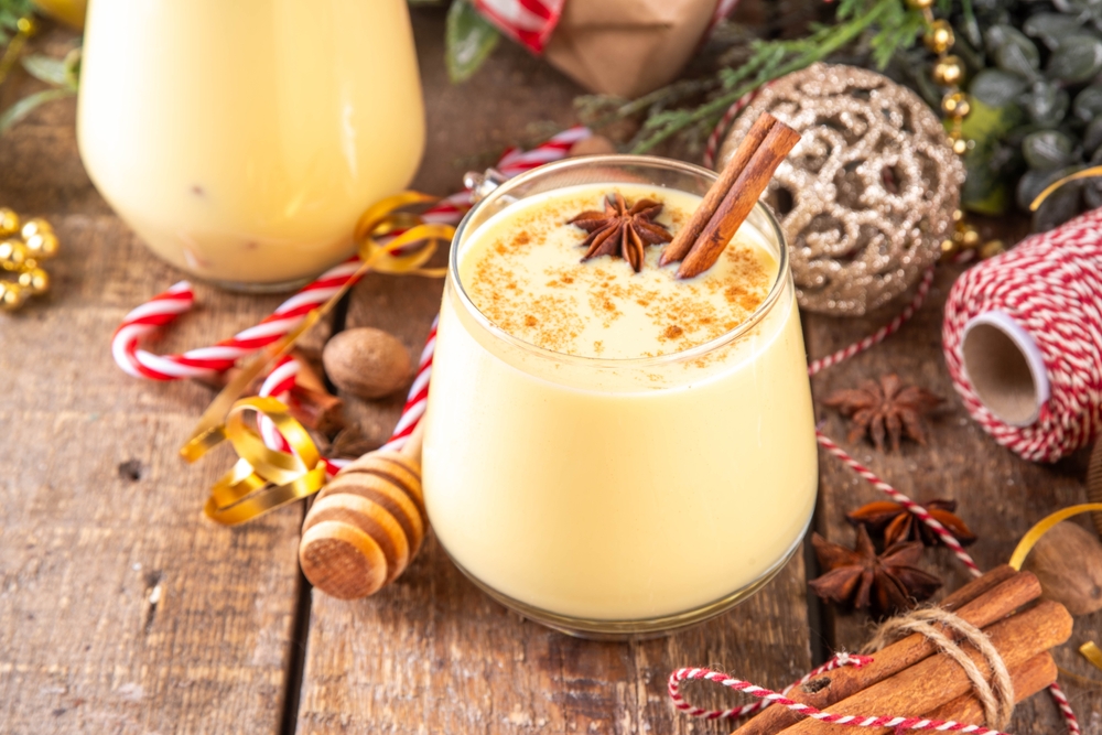 A glass of eggnog with a cinnamon stick on a table with holiday decorations.