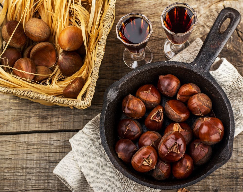 Flat-lay of roasted chestnuts in a cast iron pan next to two drink glasses filled with wine.