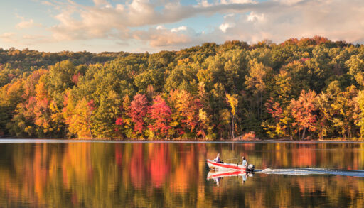 A small, red fishing boat on a glassy lake with the autumn trees reflected in its surface.