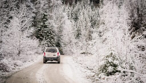 White car driving down a snow covered road lined with snowy trees.