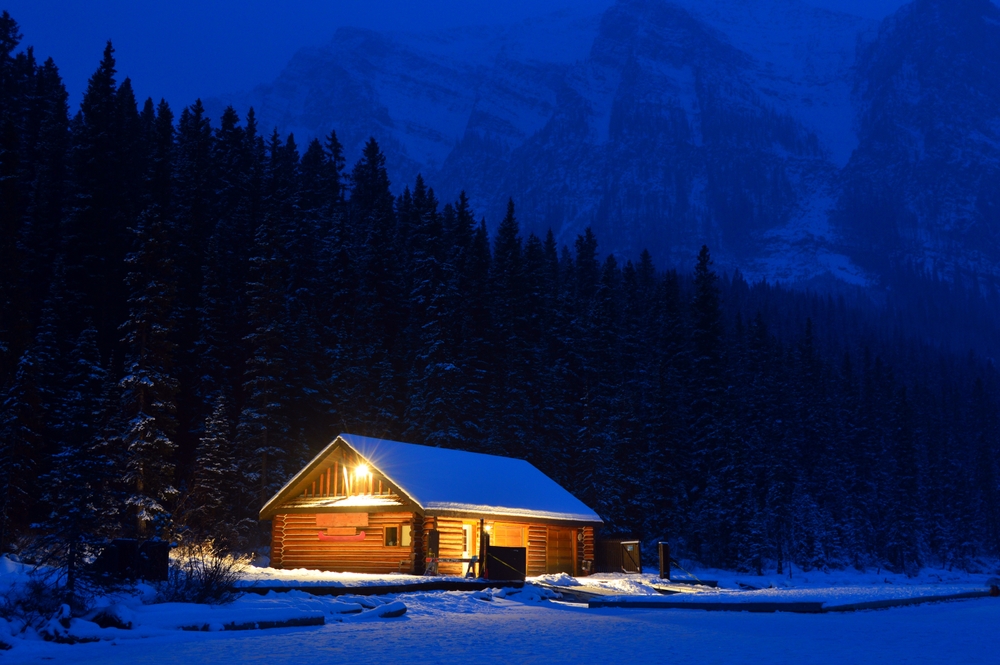 Illuminated log cabin surrounded by snow and trees at dusk in Banff National Park, Alberta.