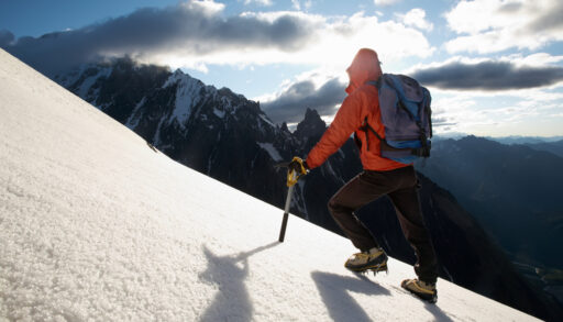 Person hiking up a mountain in an orange jacket with more mountains in the background.