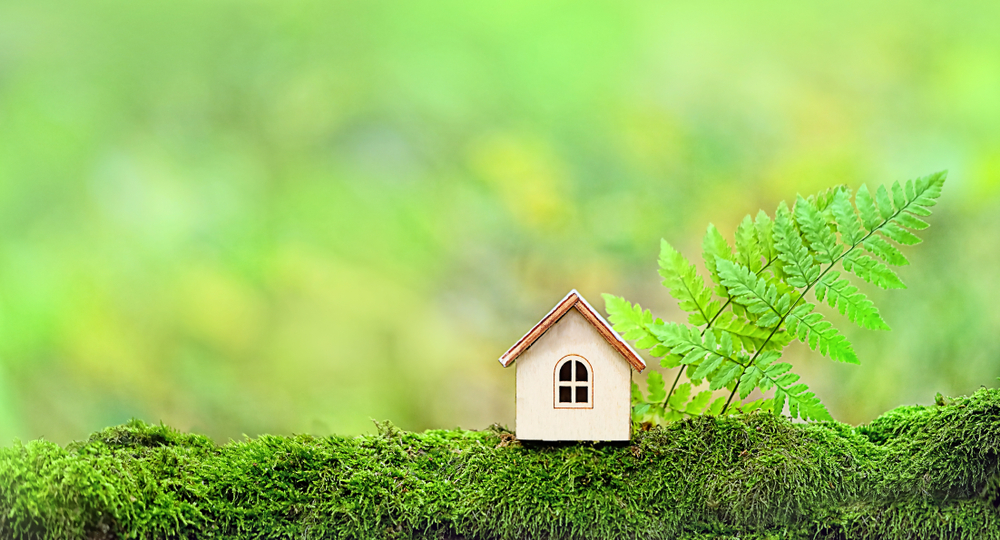 Small toy house sitting on a mossy tree branch with a green leaf next to it.