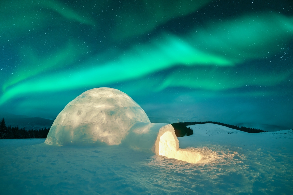 Igloo with a light coming from it against the Northern lights and mountain landscape.
