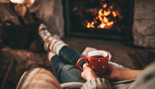 Person sitting indoors in front of a fire with a warm drink in their hands.