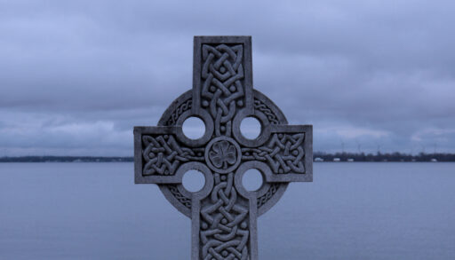 Close-up of celtic cross overlooking a body of water at dusk.