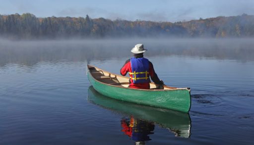 Man in a white fishing hat canoeing on a lake in a green canoe.