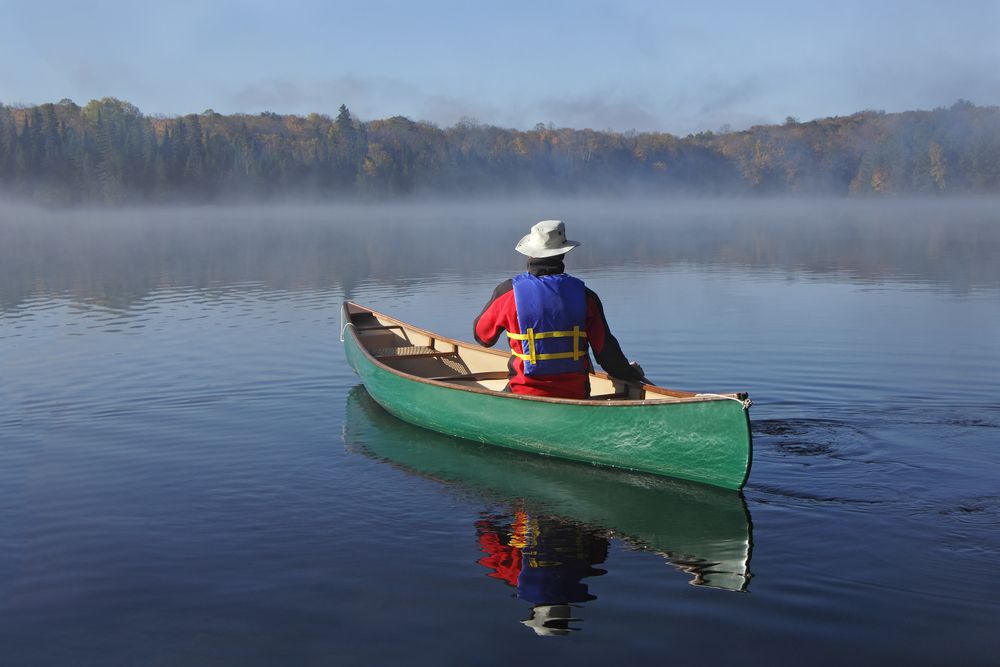 Man in a white fishing hat canoeing on a lake in a green canoe.