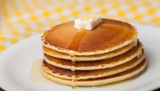 A stack of pancakes with a pad of butter and syrup on a white plate sitting on a yellow, gingham tablecloth.