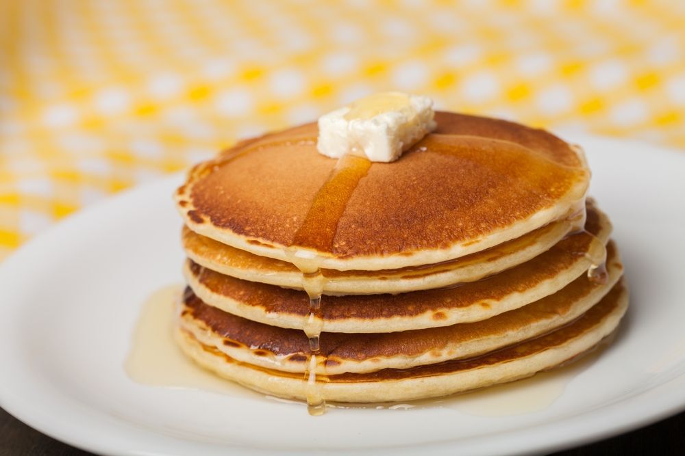 A stack of pancakes with a pad of butter and syrup on a white plate sitting on a yellow, gingham tablecloth.