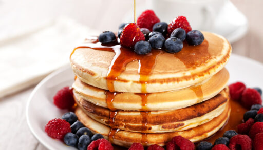 Maple syrup being poured on a stack of pancakes topped with berries.