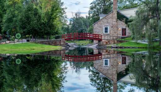 Small stone cottage with a red bridge over a clear river and a tree landscape.