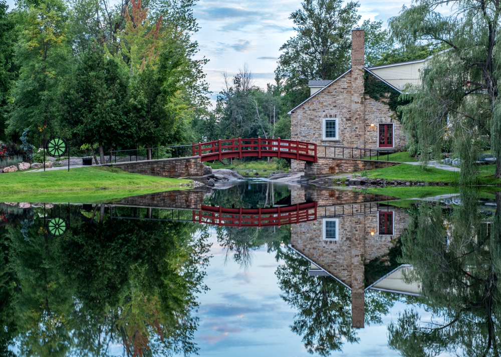 Small stone cottage with a red bridge over a clear river and a tree landscape.