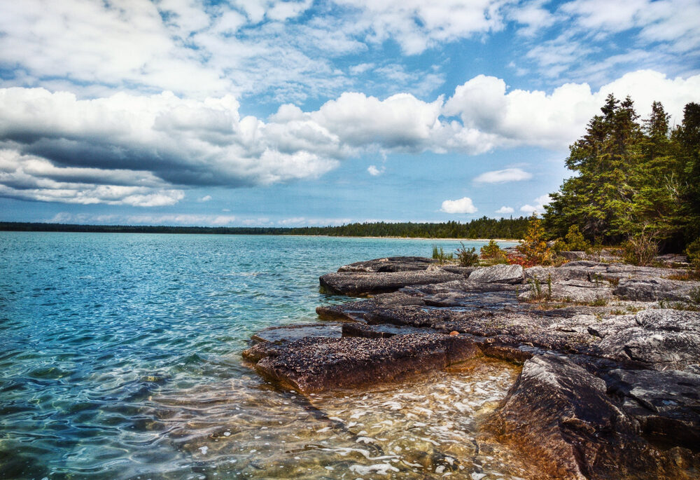 Clear waters of Lake Huron and the rocky shoreline lined with trees.