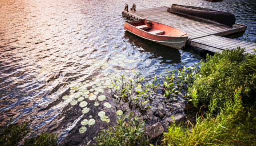 An old red rowboat anchored next to a wooden dock on a lake with the sun reflecting on it.