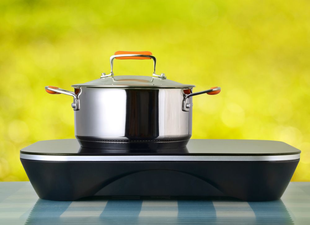 Induction cooktop with a pot on top of it on a blue gingham table against a yellow background.