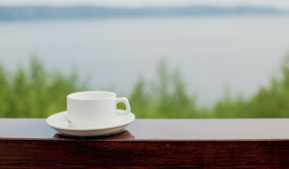 White coffee cup and saucer on a wooden deck railing overlooking a lake.