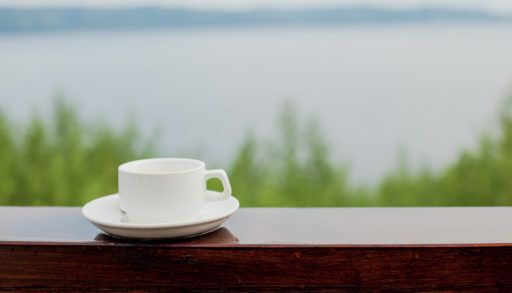 White coffee cup and saucer on a wooden deck railing overlooking a lake.