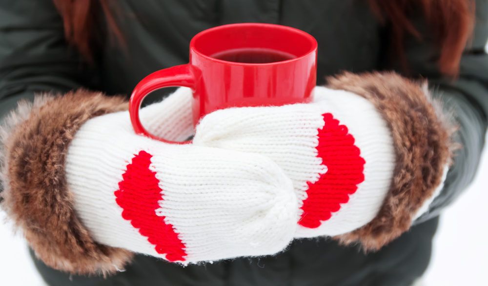 Woman wearing white mittens with read hearts on them holding a red mug of hot cocoa.