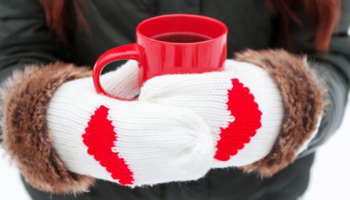 Woman wearing white mittens with read hearts on them holding a red mug of hot cocoa.