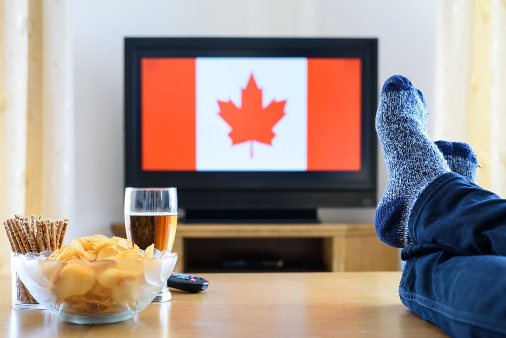 Person with their feet up and a bowl of chips and beer next to them with a Canadian flag displayed on a TV.