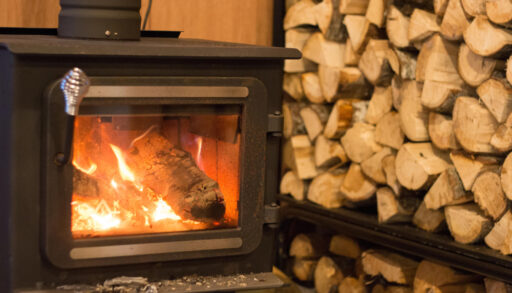 Woodstove fire burning with a pile of wood stacked in the background.