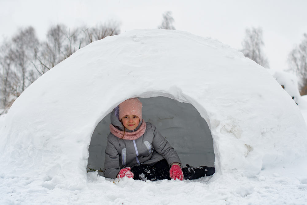 Child in a grey snowsuit laying on the ground in an igloo.