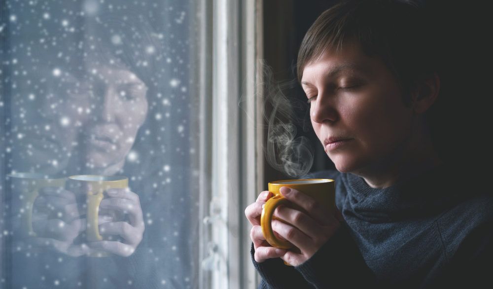 Woman looking out a window with a forlorn expression on her face and a yellow mug in her hands.