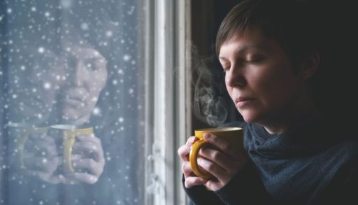 Woman looking out a window with a forlorn expression on her face and a yellow mug in her hands.