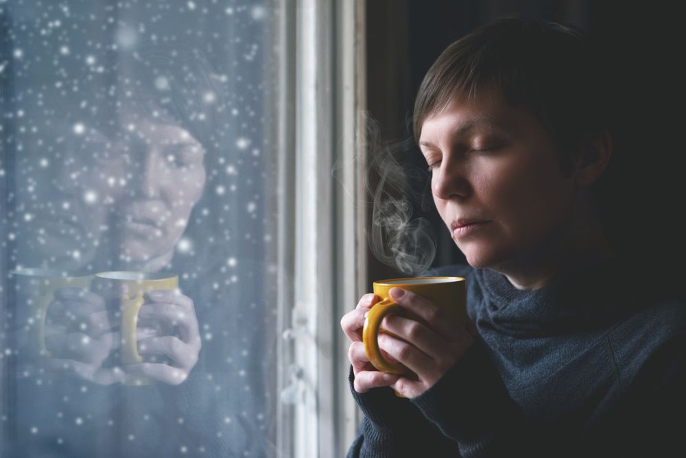 Woman looking out a window with a forlorn expression on her face and a yellow mug in her hands.
