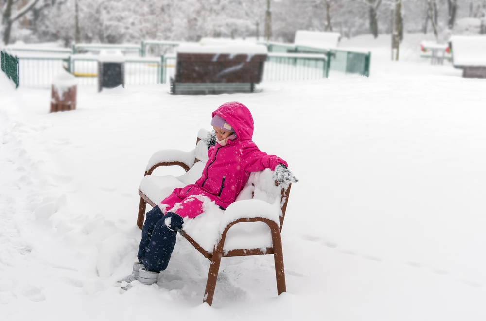 Bored little girl in pink snowsuit sitting on a bench covered in snow.