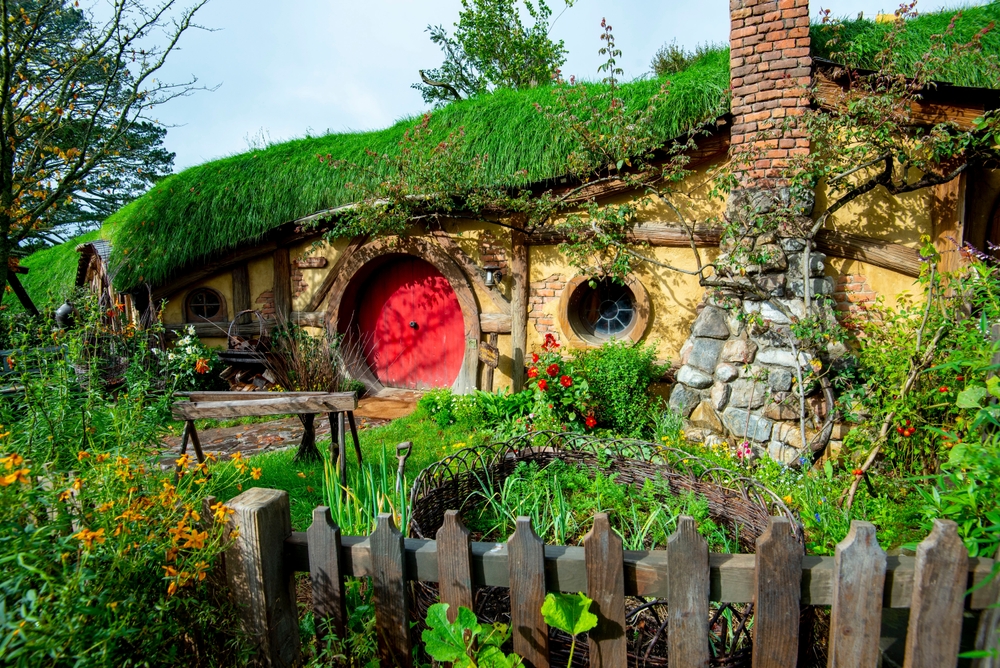 Hobbit hole with a red circular door and stone walls covered in moss and overgrowth.