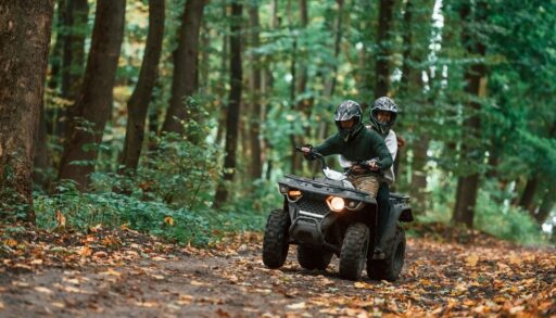 Two people riding an ATV on a trail in a thickly wooded forest.