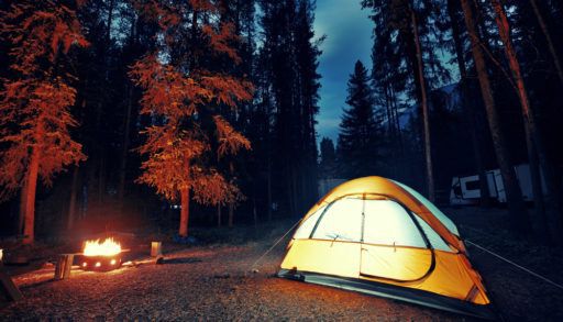 Yellow tent lit up next to a fire at dusk in the woods.