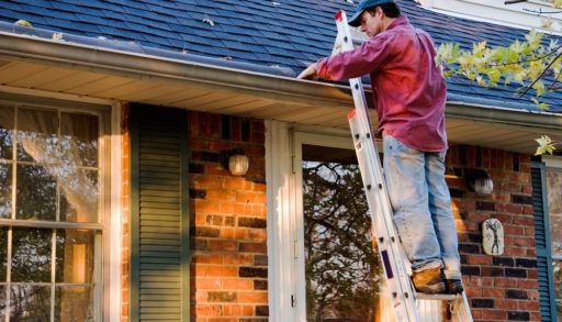 Man in a red plaid shirt and a blue hat on a ladder cleaning out the gutters of his house.