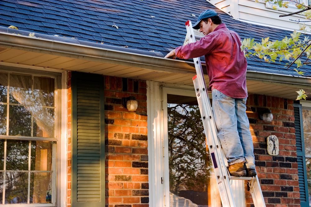 Man in a red plaid shirt and a blue hat on a ladder cleaning out the gutters of his house.